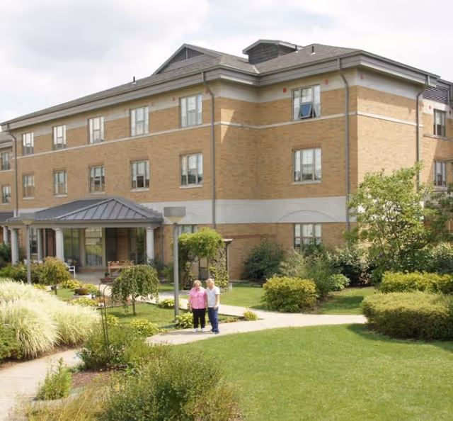 A three-story brick building with multiple windows, surrounded by a well-maintained garden with green grass, bushes, and small trees. Two elderly people are standing on a paved pathway in front of the building. The sky is partly cloudy.