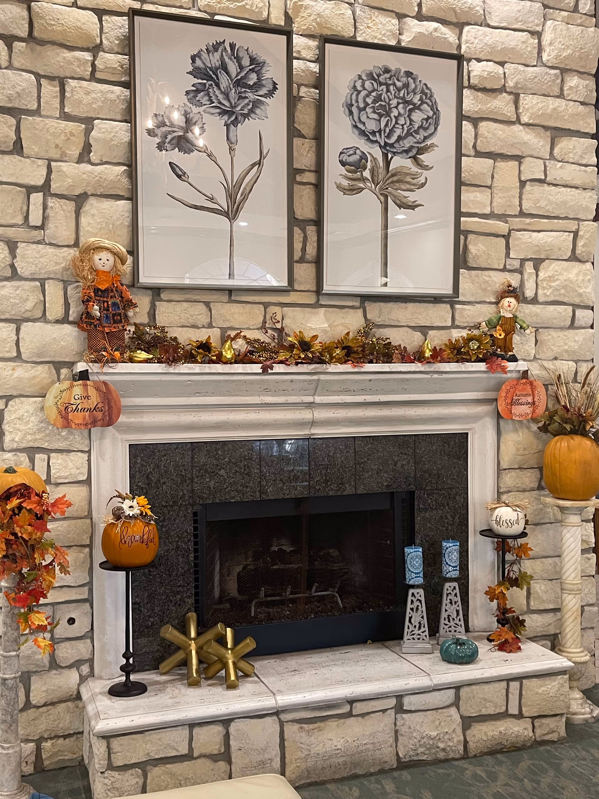Stone fireplace decorated with autumn-themed items including pumpkins, fall leaves, small scarecrow dolls, and framed botanical prints of flowers hanging above the mantel.