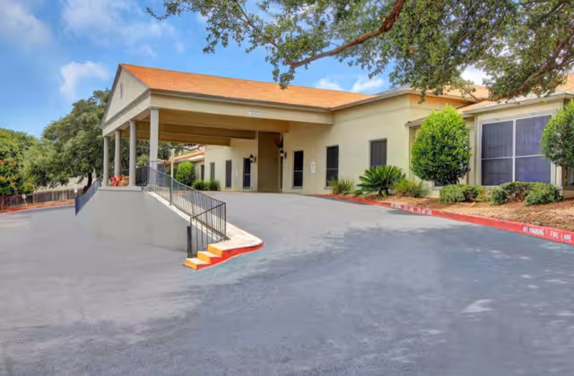 Exterior view of Wurzbach Nursing and Rehabilitation facility showing a covered entrance with a ramp and stairs, surrounded by trees and shrubs under a partly cloudy sky.