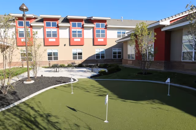 Sunny courtyard with a small putting green, patio tables, and a two-story building with red window accents.