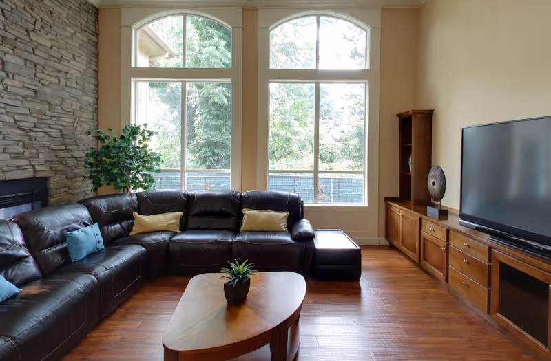 A cozy living room with a large dark brown leather sectional sofa adorned with blue and beige pillows. There is a wooden coffee table with a small plant on it in front of the sofa. The room features large windows with an outdoor view of trees, a stone accent wall with a fireplace, and a wooden entertainment unit with a flat-screen TV and decorative items.
