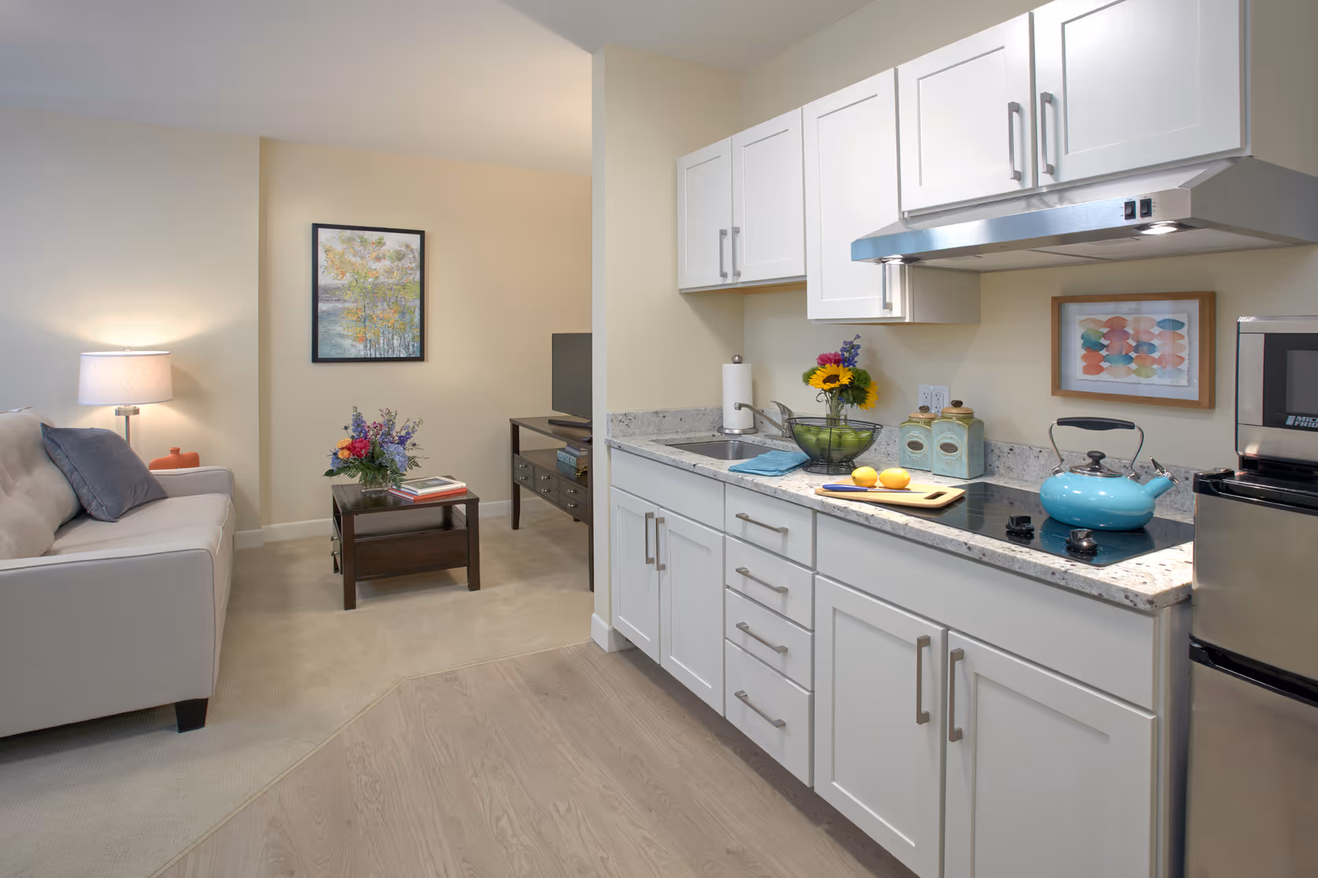 A bright and clean senior living apartment featuring a small kitchen area with white cabinets, a stovetop with a blue kettle, a sink, and a countertop with a bowl of fruit and flowers. Adjacent to the kitchen is a cozy living room with a beige sofa, a side table with a lamp, a coffee table with a flower arrangement and books, and a TV on a stand. The walls are decorated with framed artwork.