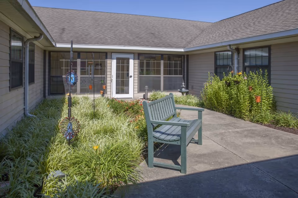 Outdoor courtyard area at Charter Senior Living of Columbia featuring a green wooden bench on a concrete pathway surrounded by lush green plants and flowers. The courtyard is enclosed by beige siding walls with multiple windows and a white door leading inside. Decorative hanging ornaments are placed among the plants.