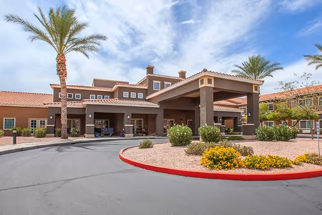Exterior view of a senior living facility with a covered entrance, palm trees, and landscaped areas with bushes and yellow flowers under a partly cloudy sky.
