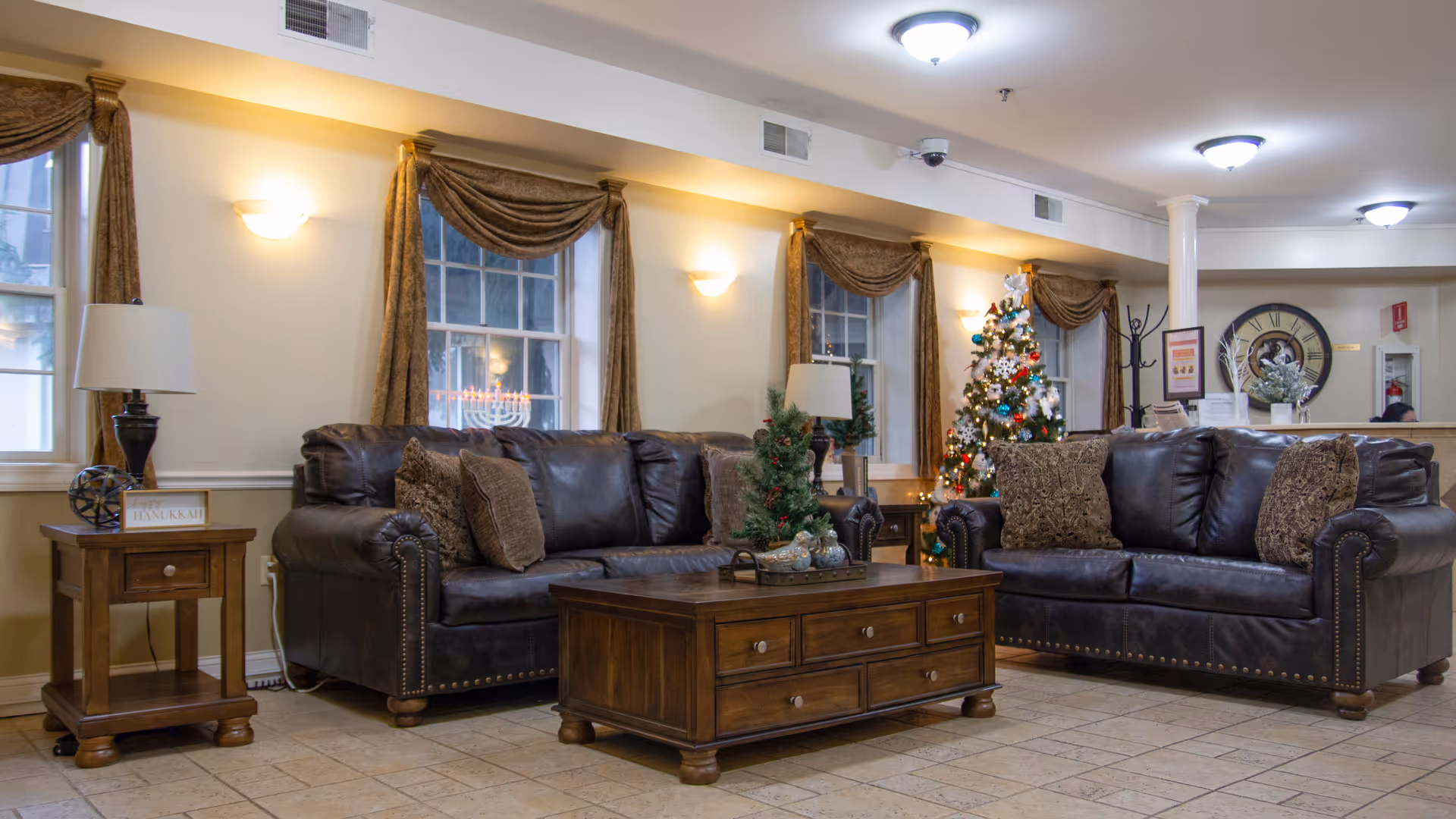 A cozy living room area in Lincoln Park Care Center featuring two dark brown leather sofas with decorative pillows, a wooden coffee table with holiday decorations, a side table with a lamp and a menorah, and a decorated Christmas tree near the back wall. The room has beige walls, windows with brown curtains, and ceiling lights.