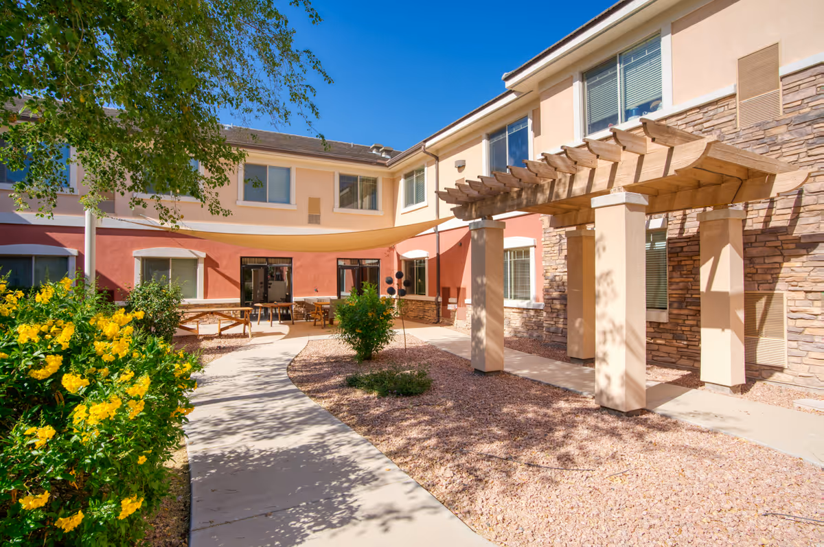 Sunny courtyard of a senior living facility with a shaded pergola, walkway, seating area and flowering bushes in front of a two-story building.