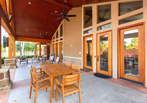 Covered outdoor patio area with wooden ceiling and ceiling fans, featuring a wooden dining table with six chairs in the foreground and additional seating with cushions in the background. Large glass doors with wooden frames lead into the building, and greenery is visible beyond the patio.