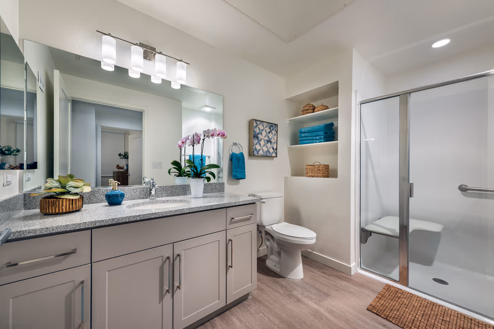 A modern bathroom featuring a large mirror with four-light fixture above a granite countertop with a sink. The countertop has decorative plants and a soap dispenser. There is a toilet next to built-in shelves holding folded towels and baskets. A glass-enclosed shower with a built-in bench is visible on the right side. The floor has wood-like tiles and a small woven mat is placed in front of the shower.