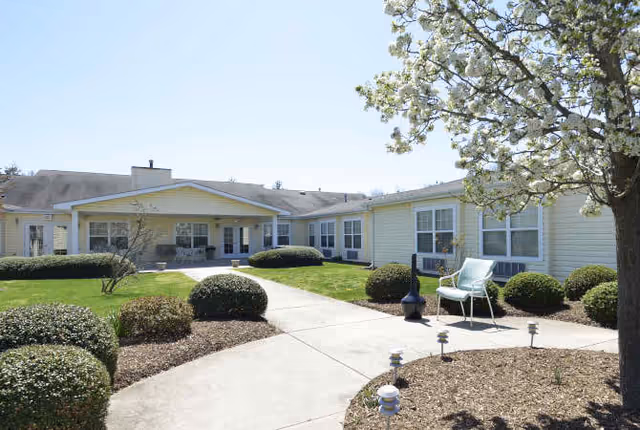 Exterior view of a single-story senior living facility building with light yellow siding, multiple windows, and a covered entrance. The foreground features a concrete walkway surrounded by landscaped bushes, mulch beds, a flowering tree, and a blue chair on the right side.