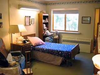 A furnished senior living bedroom with a blue-patterned bedspread, window, lamp, and bookshelf.