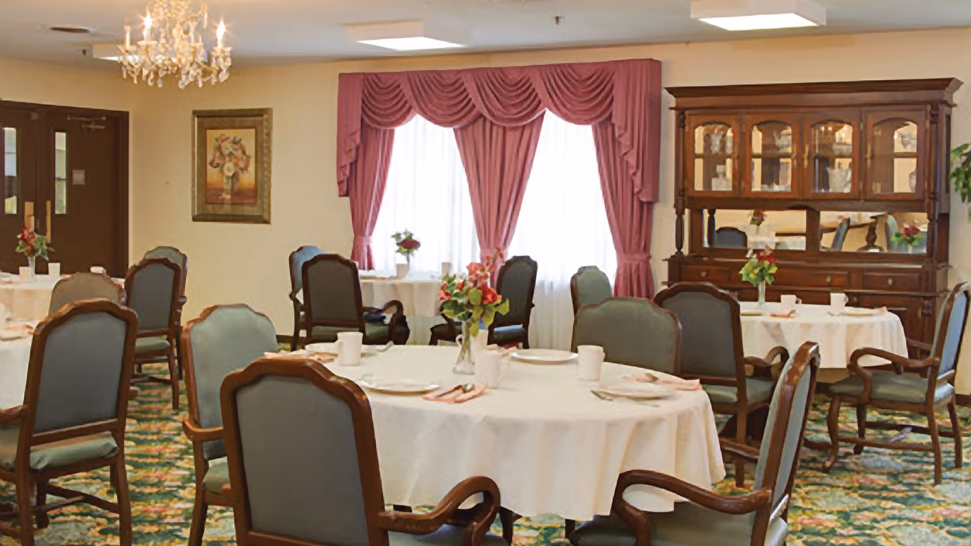 A dining room with round tables covered in white tablecloths, each set with plates, cups, and silverware. The chairs have wooden frames with green upholstery. There are floral centerpieces on each table. The room features a chandelier, a large window with pink drapes, a wooden china cabinet, and a framed painting on the wall.