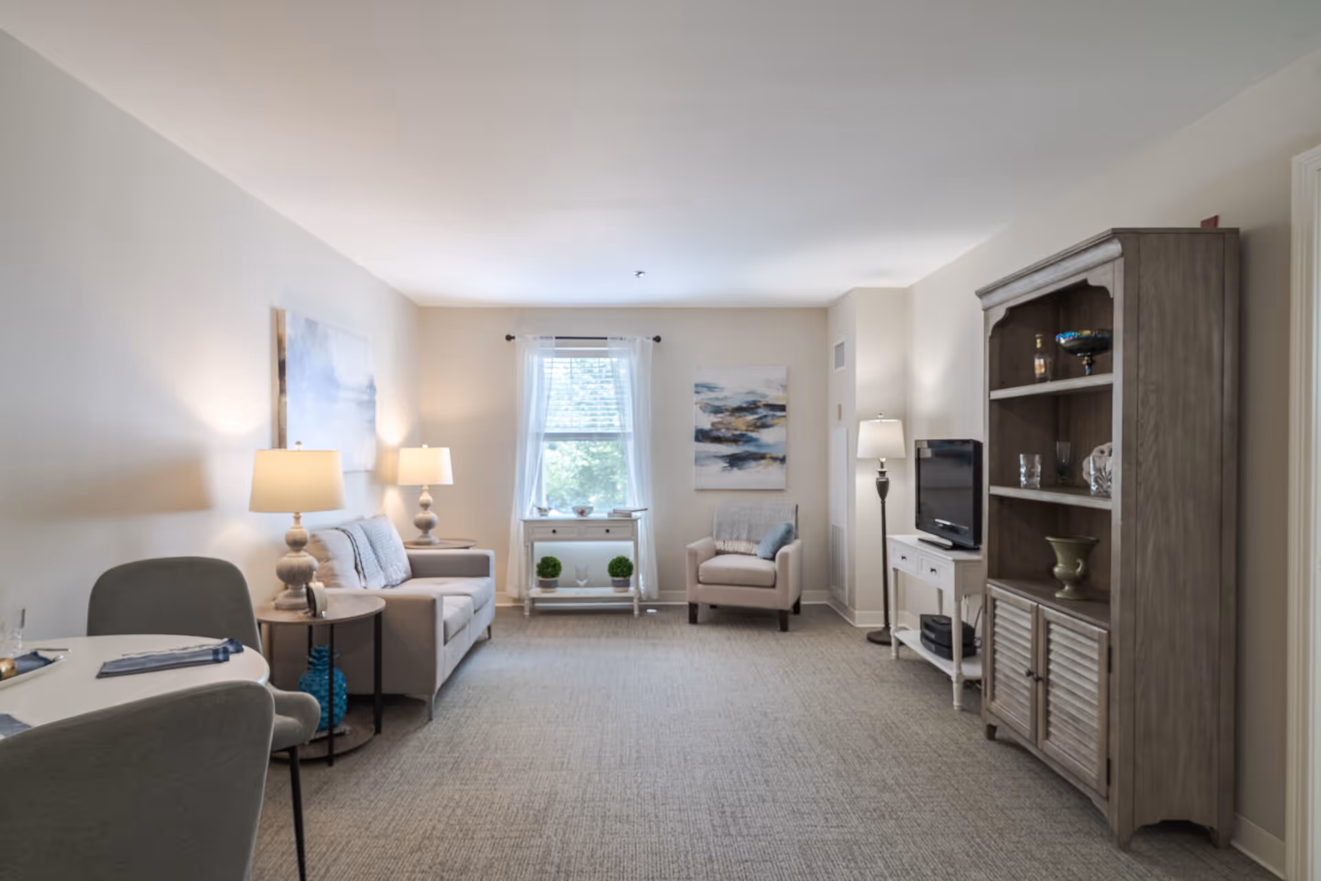 A bright and cozy living room with neutral tones featuring a gray sofa with cushions, a matching armchair, two table lamps on side tables, a window with sheer white curtains, a white console table with decorative plants, a floor lamp, a flat-screen TV on a white stand, and a large wooden cabinet with shelves displaying decorative items.