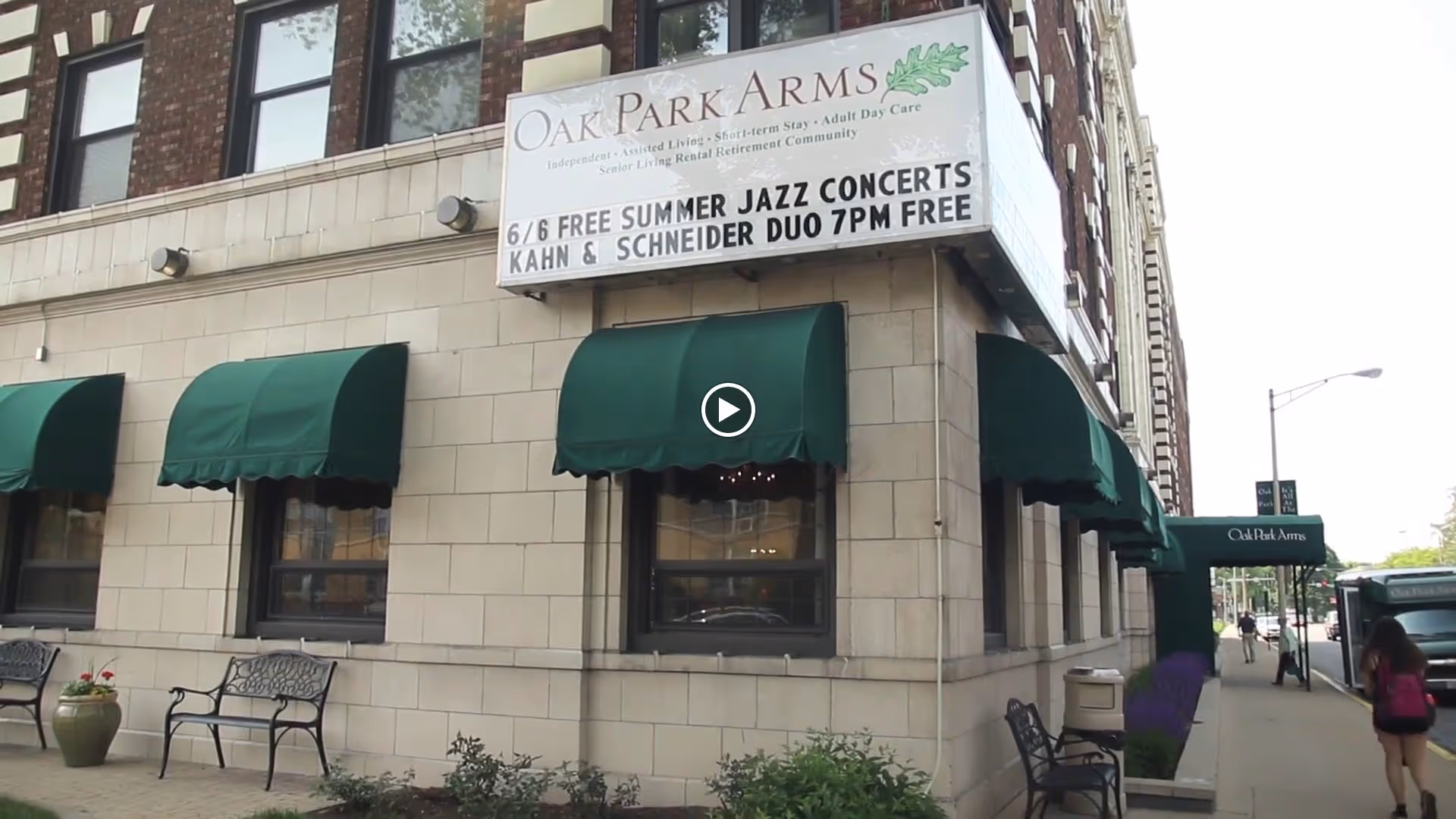 Exterior corner view of Oak Park Arms building with green awnings over windows and entrance. A sign above the corner advertises free summer jazz concerts featuring Kahn & Schneider Duo at 7 PM. Benches and planters are placed along the sidewalk.