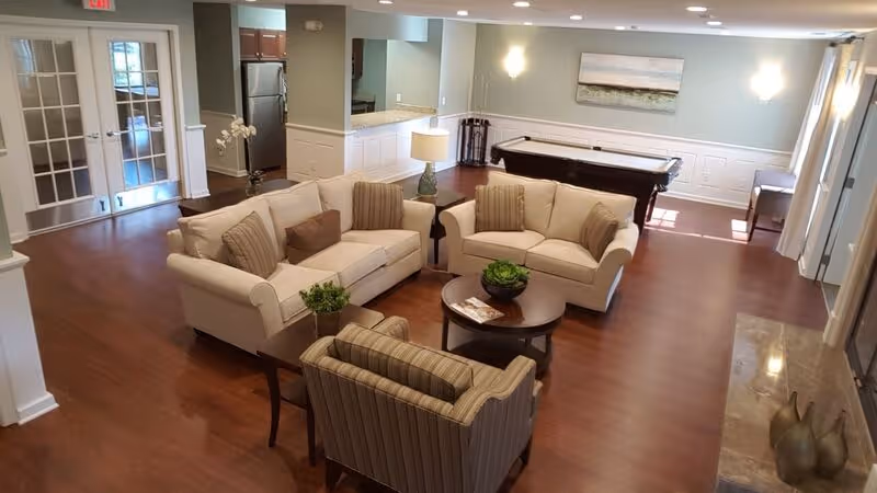 Open communal lounge with sofas, armchairs, a coffee table, a pool table and a small kitchenette in a senior apartment common area.