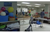 Physical therapy room with exercise equipment including parallel bars, therapy balls, and a stationary bike. Two mannequins are positioned near the therapy area, and a person is seen in the background. The room is well-lit with white walls and ceiling lights.