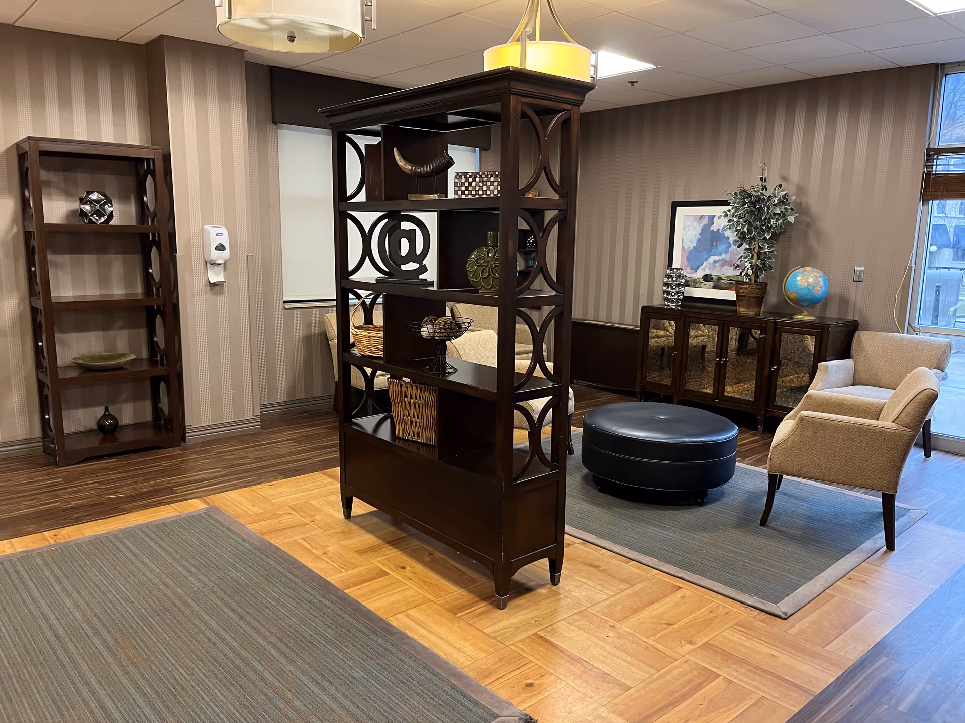 A cozy living room area with beige armchairs, a round black ottoman, and a wooden shelving unit with decorative items. The room has wood and carpet flooring, striped wallpaper, a window with blinds, and a sideboard with a globe, plant, and framed artwork.