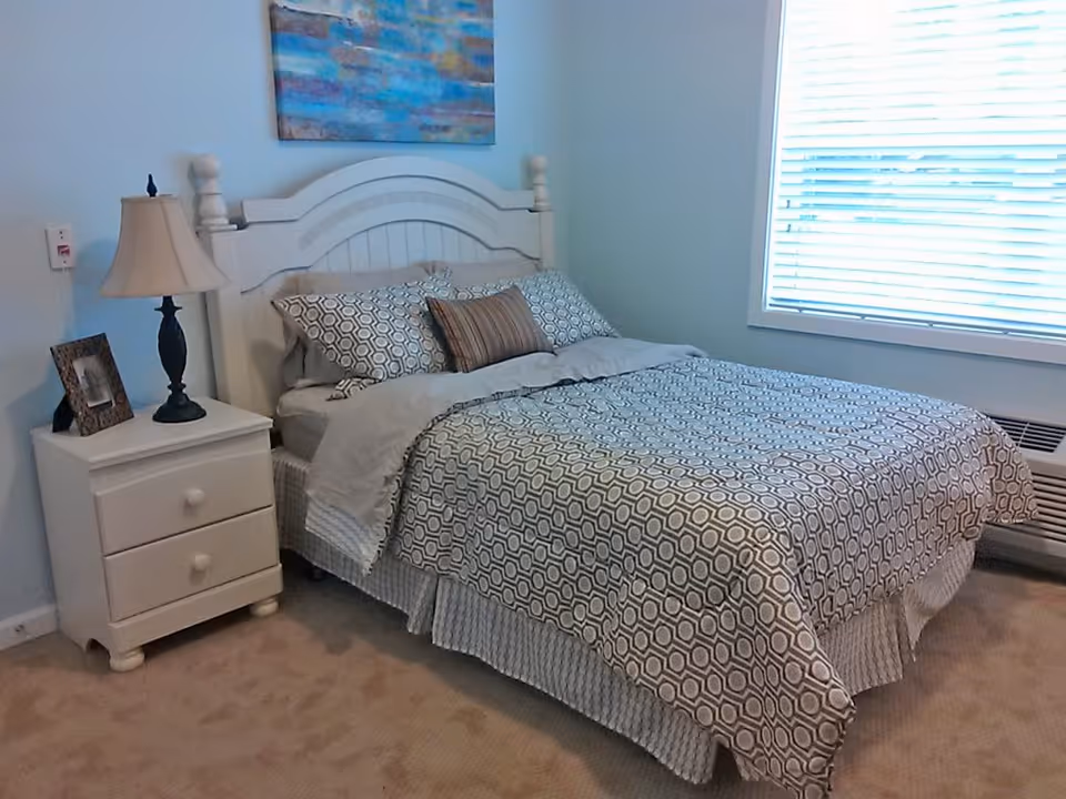 A neatly made bed with patterned bedding and pillows in a bedroom. Next to the bed is a white nightstand with two drawers, a table lamp, and a framed photo. A large window with blinds is on the right wall, and a colorful abstract painting hangs above the bed's white headboard.