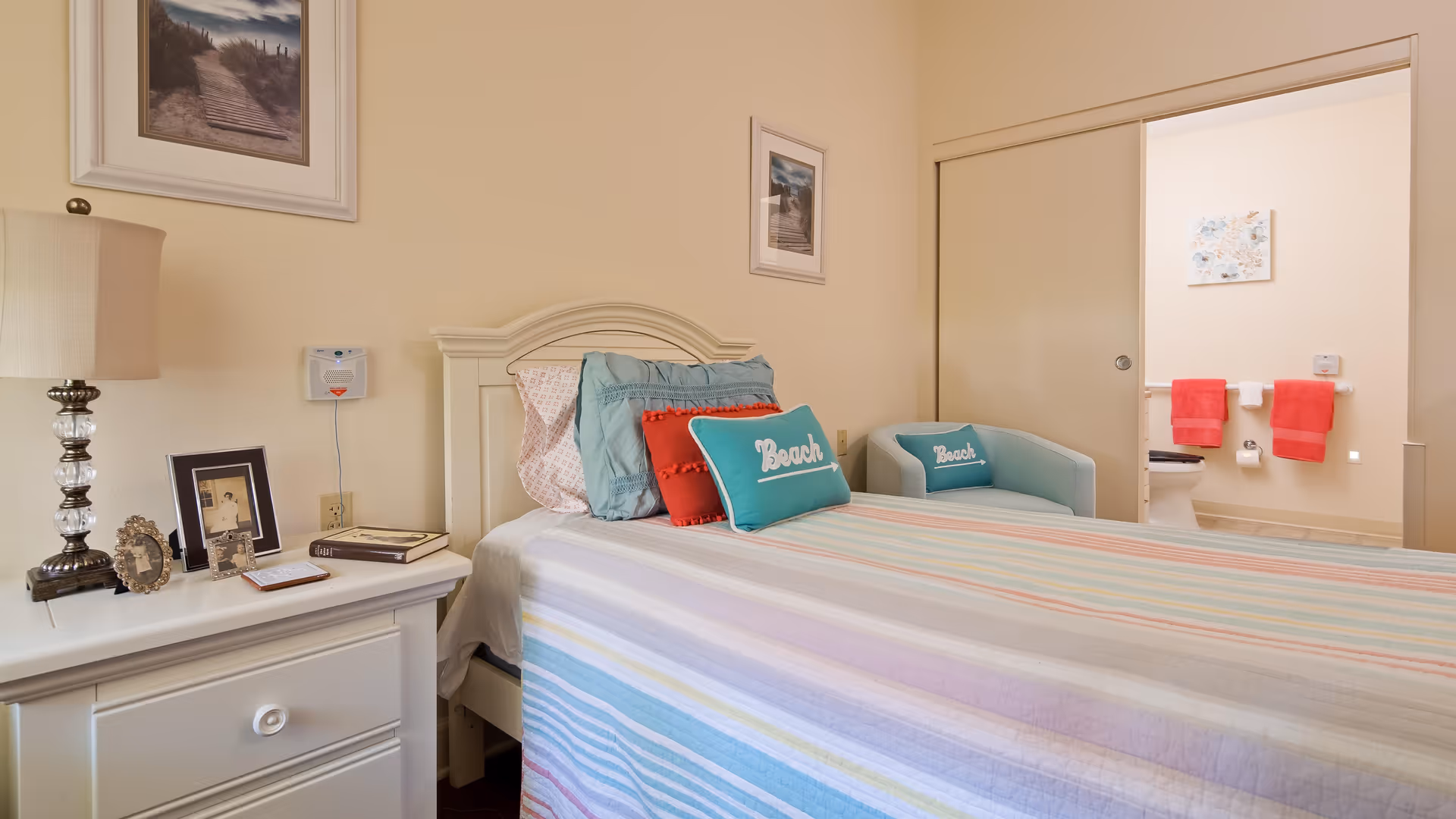 A cozy bedroom with a bed covered in a striped pastel bedspread and multiple pillows, including two with the word 'Beach' on them. There is a white nightstand with a lamp, framed photos, and books. In the background, a bathroom with red towels hanging on the wall is visible through an open sliding door.
