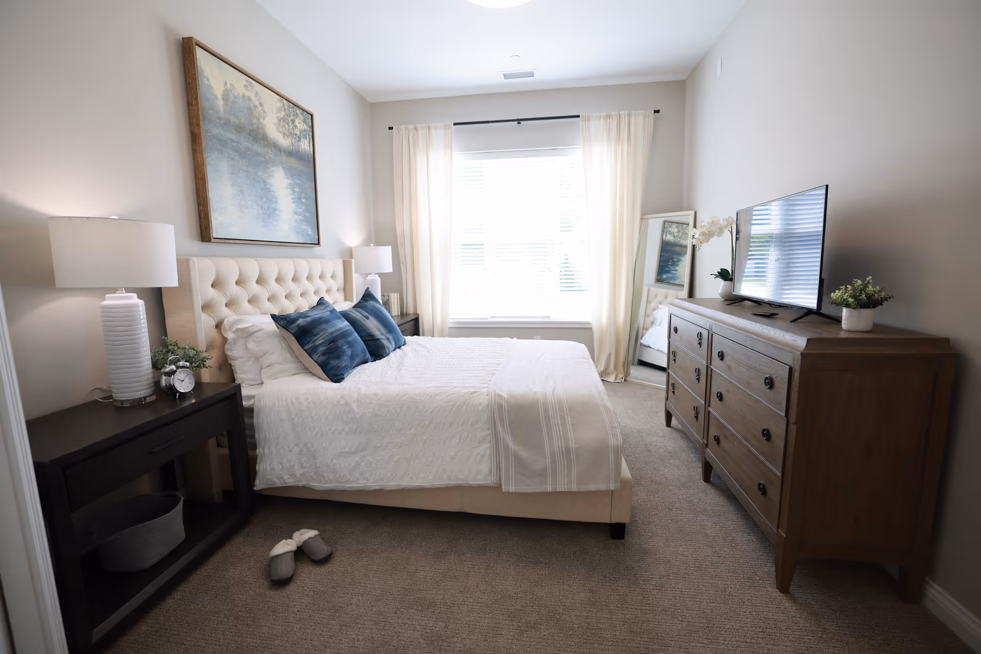 A bright and cozy bedroom featuring a beige upholstered bed with white bedding and two blue accent pillows. On the left side, there is a dark wooden nightstand with a white lamp, a small plant, and an alarm clock. A pair of gray slippers is placed on the carpeted floor near the bed. On the right side, a wooden dresser holds a flat-screen TV and a small potted plant. A large window with sheer white curtains lets in natural light, and a full-length mirror leans against the wall near the window. A framed landscape painting hangs above the bed.