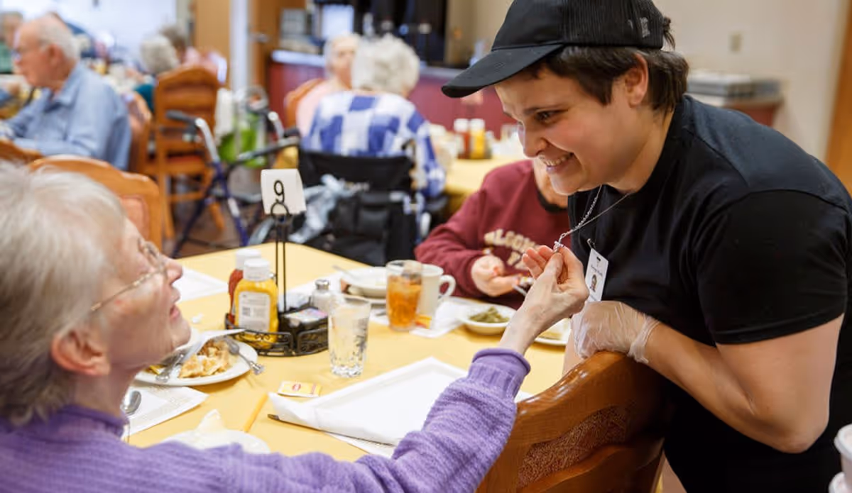 A staff member engaging with a resident at a dining table in Vintage Knolls, with food and drinks on the table and other residents in the background.