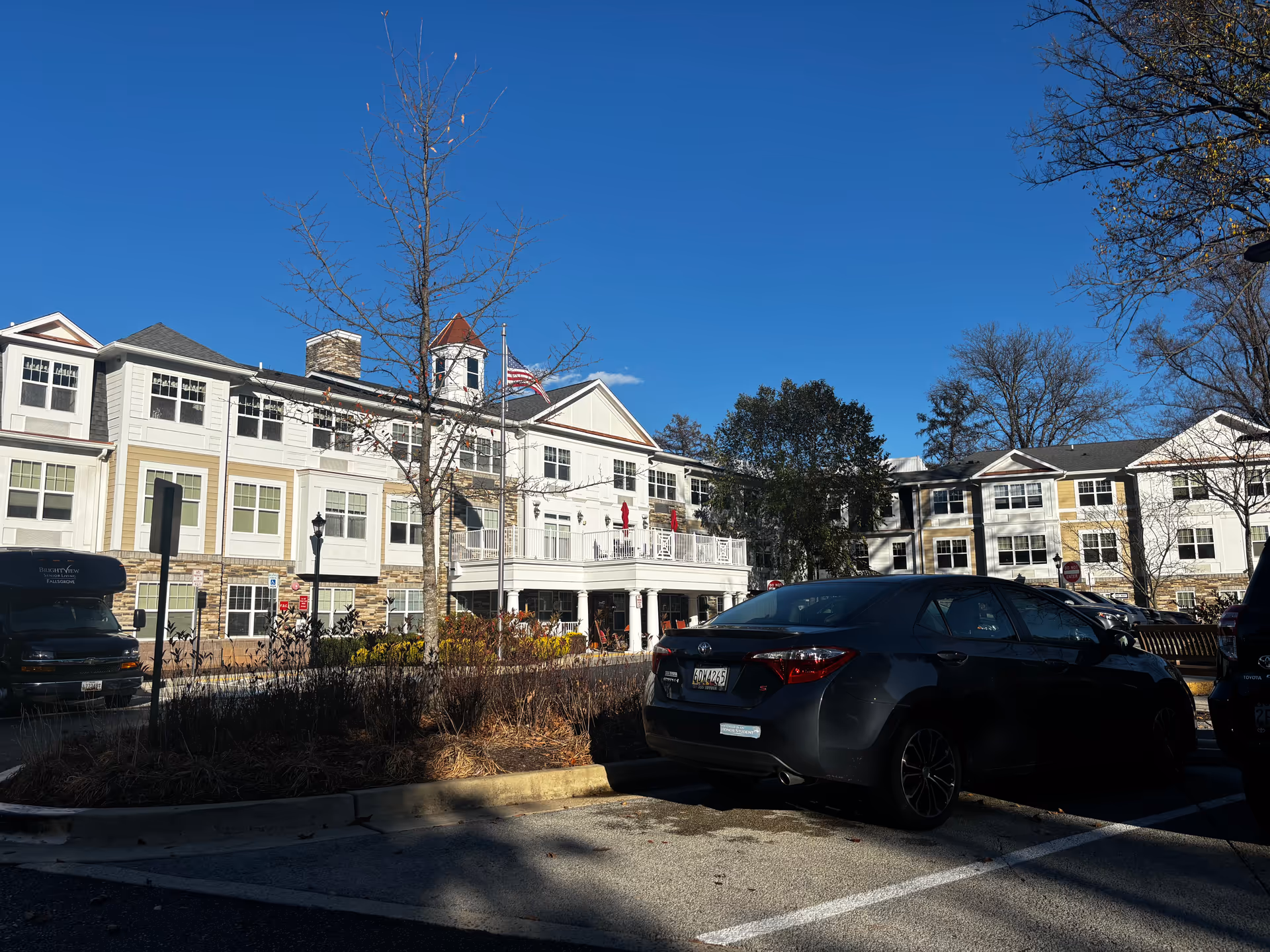 Exterior view of Brightview Fallsgrove senior assisted living and memory care facility on a clear sunny day, showing a multi-story building with white and beige siding, multiple windows, a balcony with red umbrellas, an American flag on a flagpole, and a parking lot with several cars in the foreground.