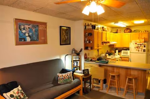 Cozy open-plan living area with sofa, wall art, ceiling fan, and a kitchen with stools
