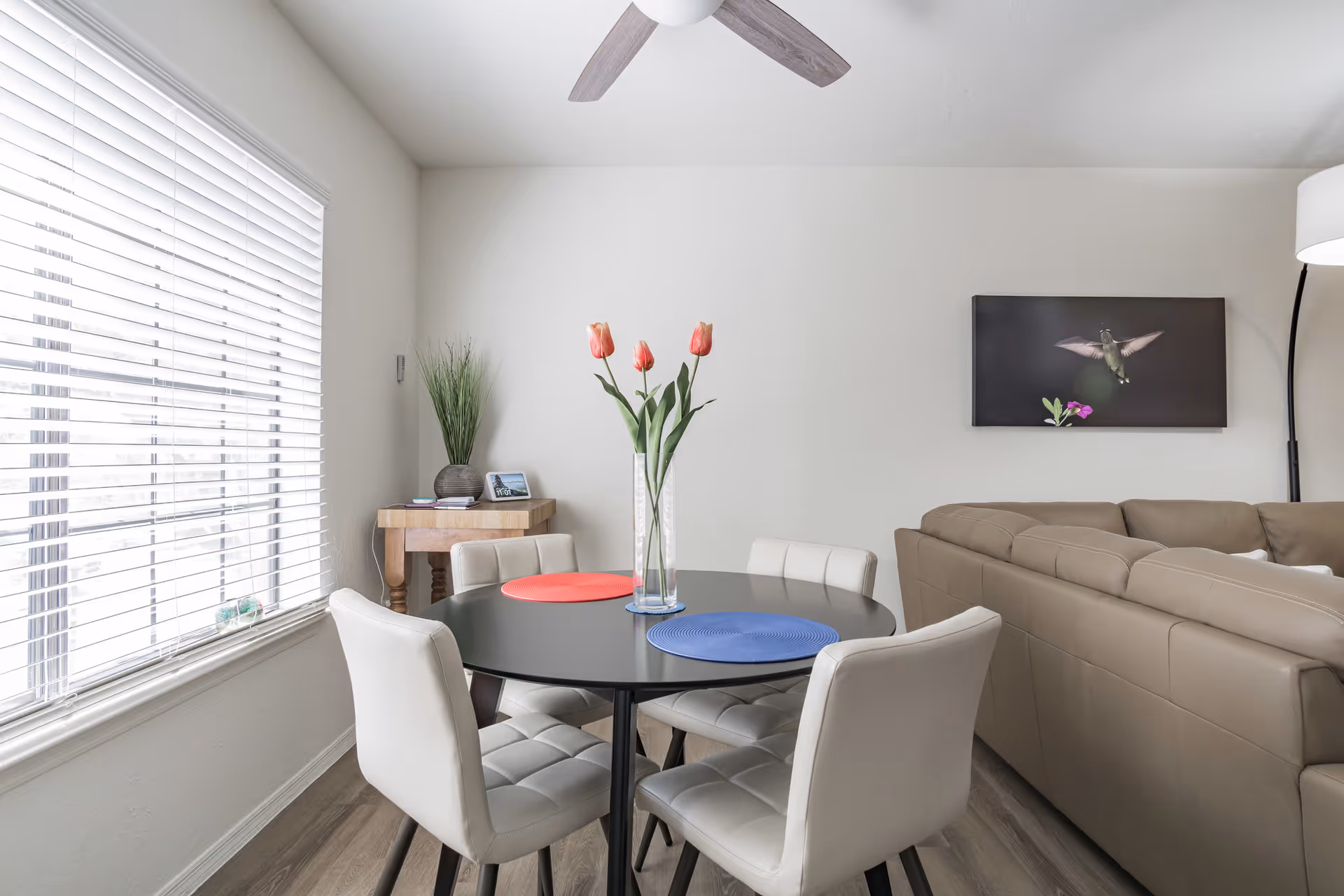 Bright dining area with a round black table, four white chairs, a vase of tulips, and a beige sectional sofa against a light wall.