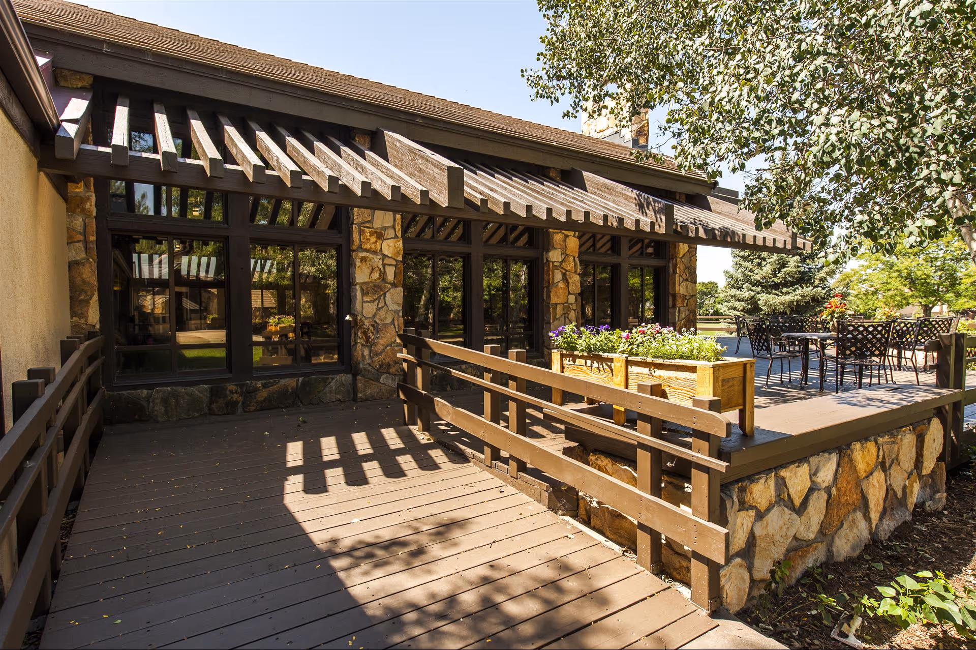 Outdoor patio area at The Gardens at Collinwood Assisted Living and Memory Care featuring a wooden deck with a railing, stone pillars, large windows, a wooden pergola, a planter box with flowers, and metal patio tables and chairs surrounded by trees and greenery.