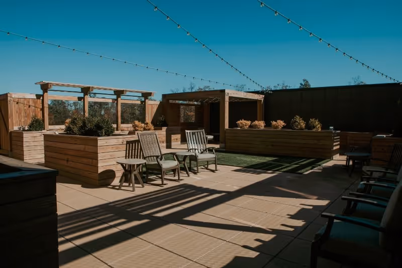 Outdoor patio area with wooden planters, chairs, small tables, string lights overhead, and a pergola under a clear blue sky.