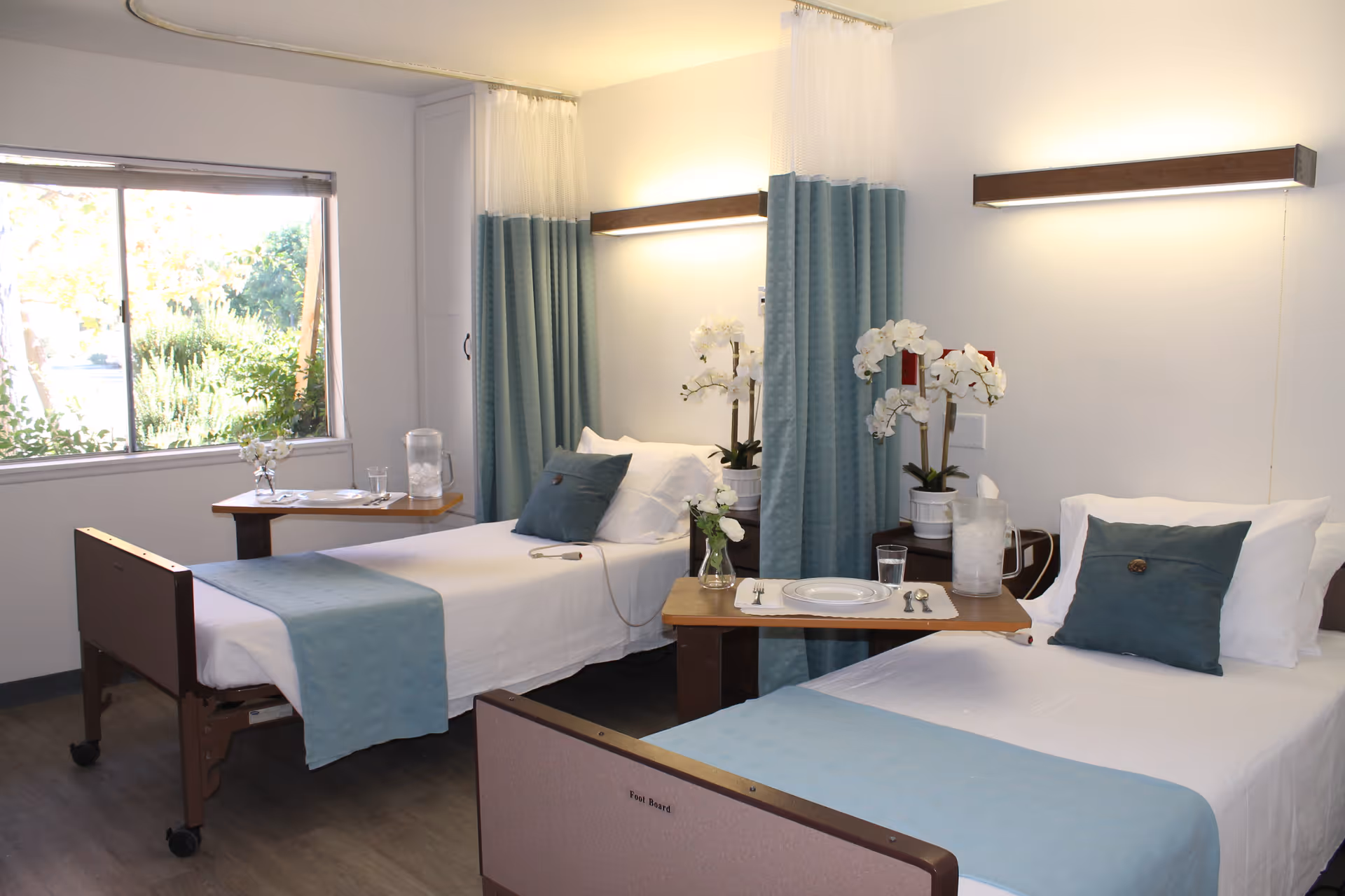 A bright room with two hospital-style beds, each with white sheets, a blue throw blanket, and a blue pillow. Between the beds are two small tables with white orchids, a glass of water, and a pitcher. Blue privacy curtains hang behind the beds, and a large window on the left side lets in natural light, showing greenery outside.