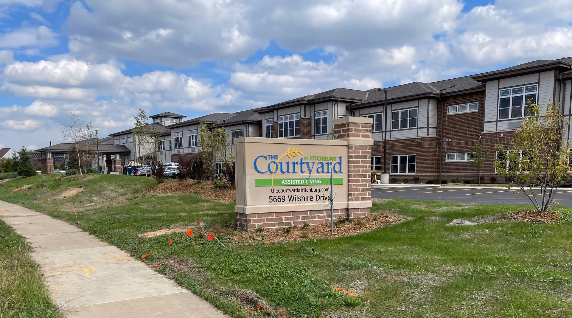 Exterior view of The Courtyard at Fitchburg assisted living building with a brick entrance sign on a grassy lawn under a partly cloudy sky.
