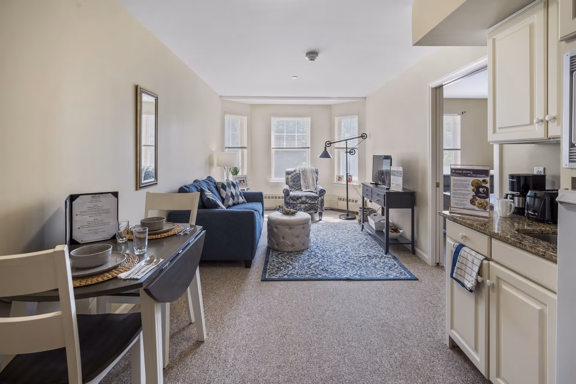 A cozy living area in a senior living facility featuring a small dining table set for two on the left, a blue sofa with patterned pillows, a patterned armchair near three windows, a blue patterned rug, a round tufted ottoman, a floor lamp, and a TV on a black console table. To the right is a compact kitchen area with white cabinets, a granite countertop, a coffee maker, and a microwave.
