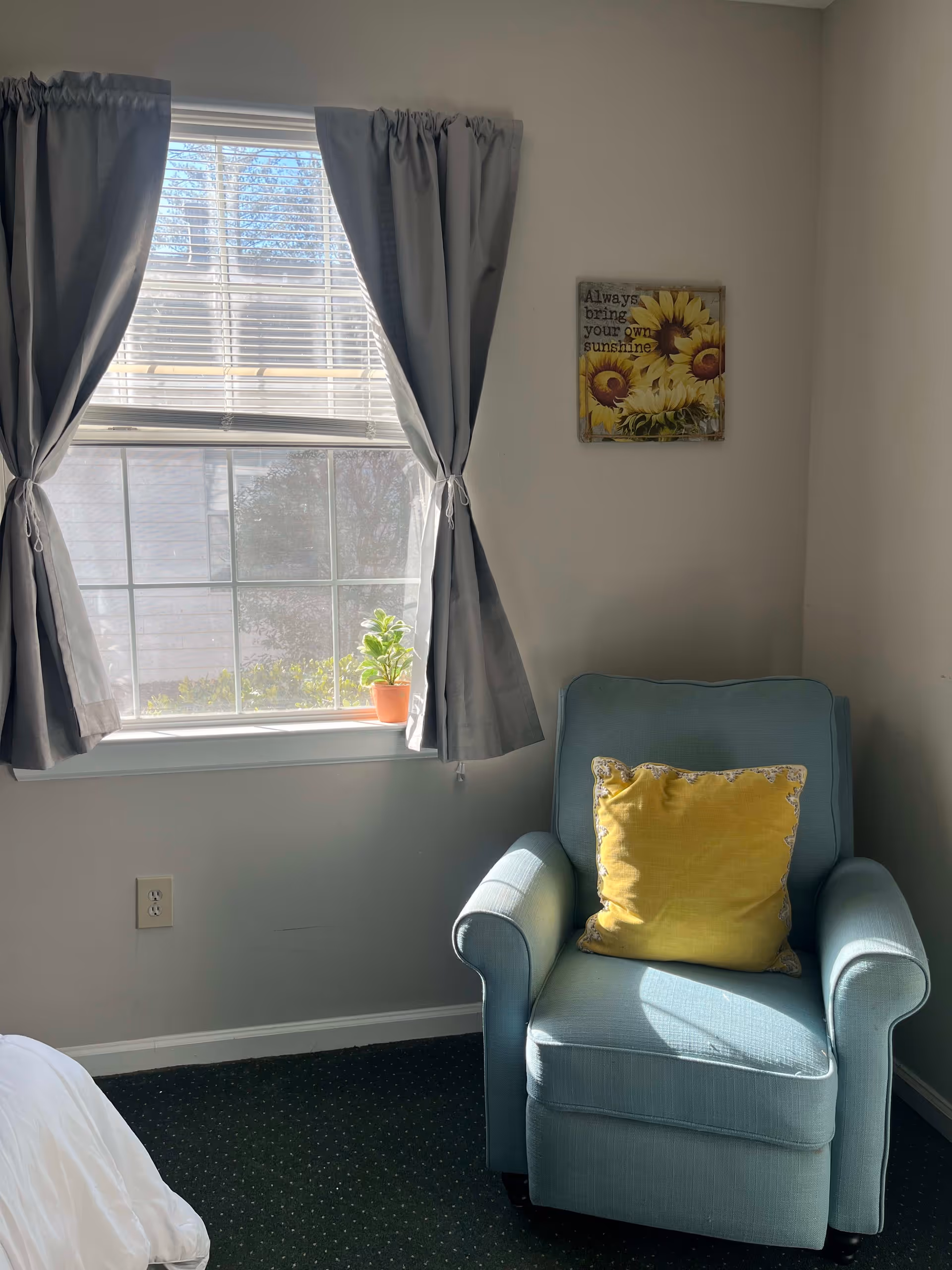 A cozy corner of a room featuring a light blue armchair with a yellow pillow, next to a window with gray curtains tied back. A small potted plant sits on the windowsill. On the wall above the chair is a decorative sign with sunflowers and the text 'Always bring your own sunshine.'