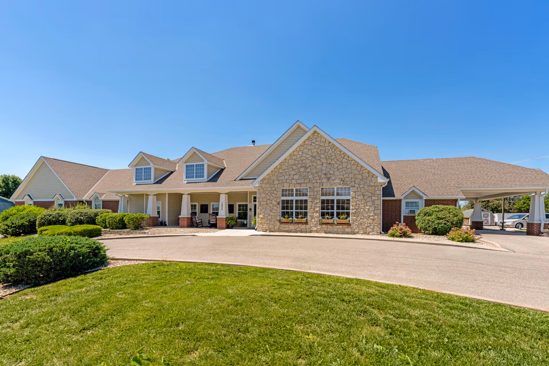 Exterior view of a single-story senior living facility building with a stone and brick facade, a covered entrance with rocking chairs, well-maintained landscaping, and a clear blue sky.