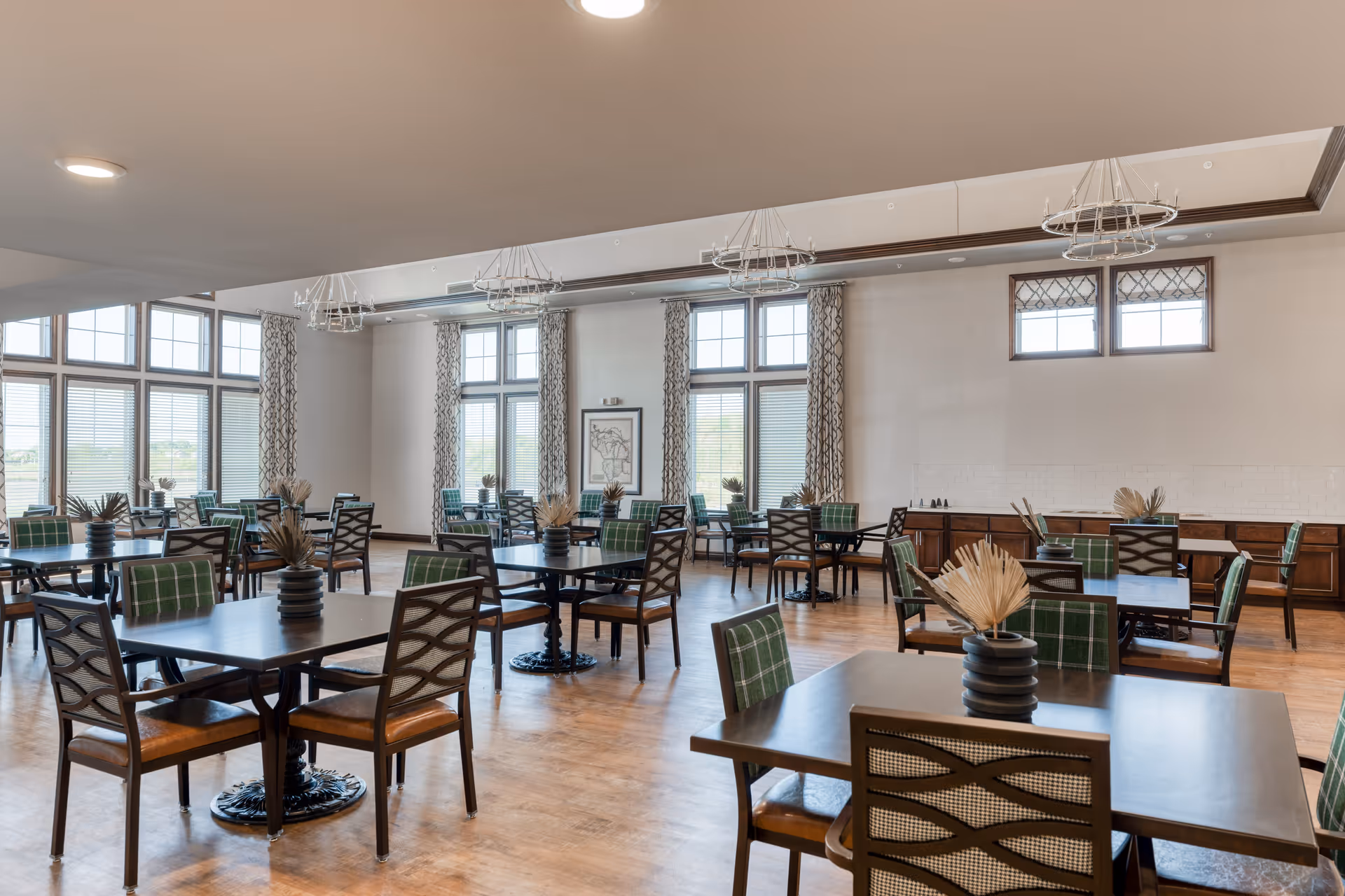A spacious dining room with multiple dark wood tables and chairs featuring green and brown cushions. The room has large windows with patterned curtains allowing natural light to fill the space. Decorative light fixtures hang from the ceiling, and each table has a small vase with dried plants.