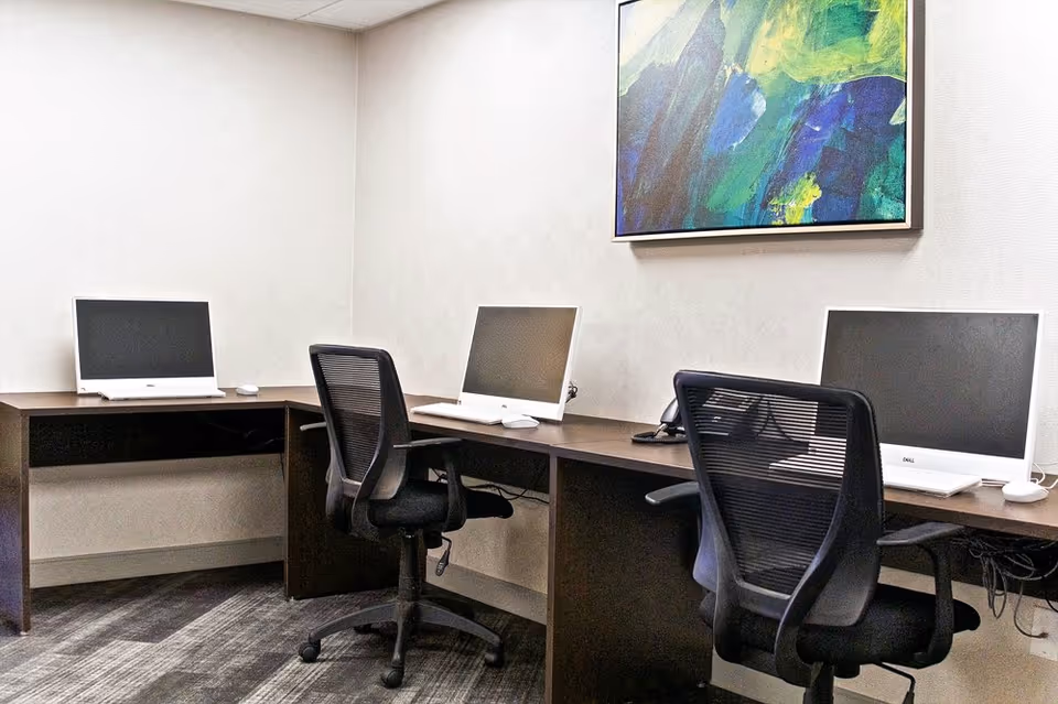 A small office or computer room with three desktop computers on a dark wooden L-shaped desk. Two black mesh office chairs are positioned in front of the computers. A colorful abstract painting hangs on the wall above the desk. The room has light-colored walls and a carpeted floor.