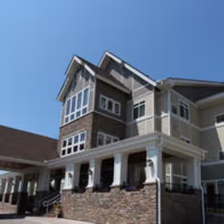 Exterior view of a multi-story assisted living facility building with stone and beige siding, large windows, and a covered entrance under a clear blue sky.