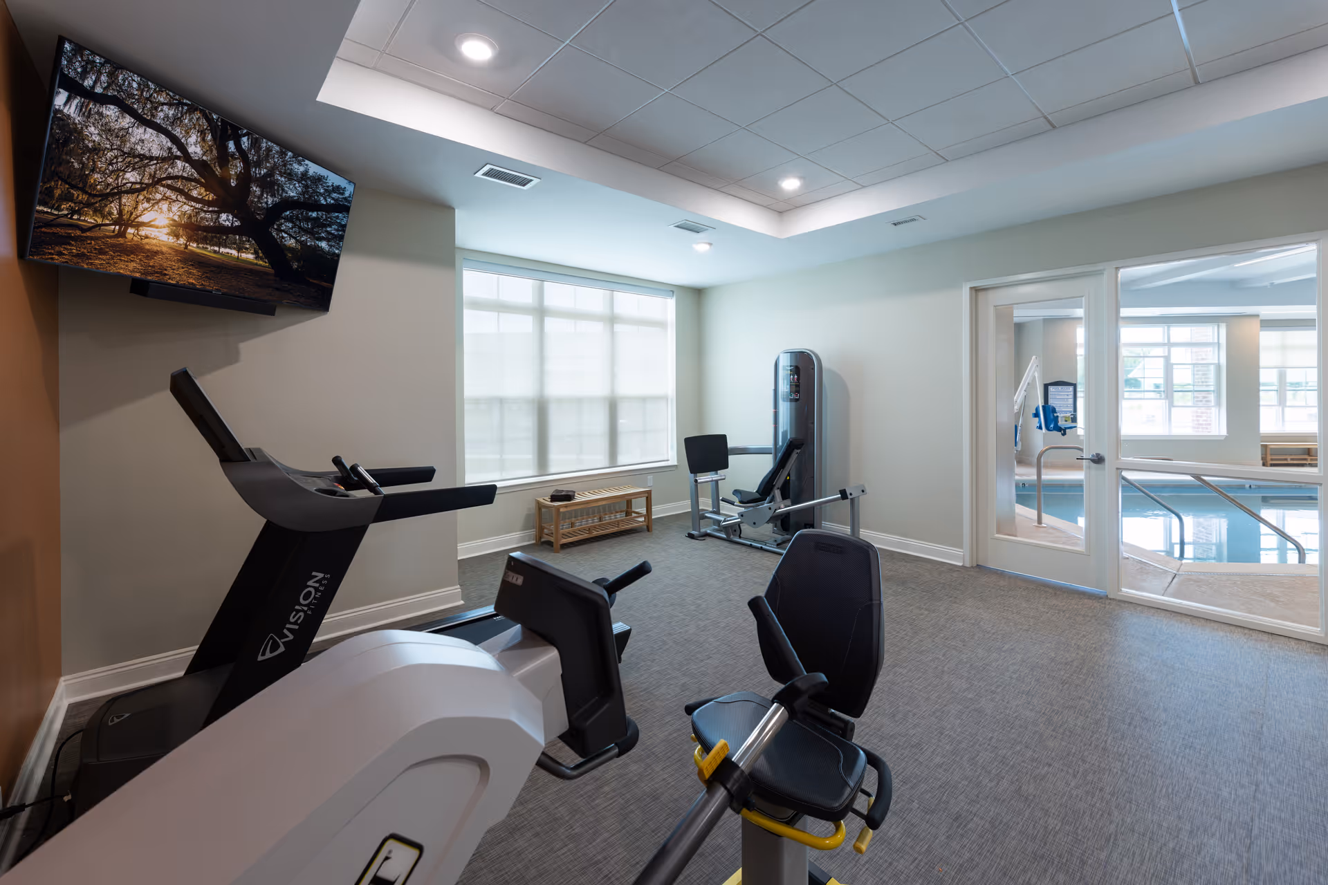 A fitness room with exercise equipment including a treadmill, a recumbent bike, and a weight machine. There is a large window with blinds, a wall-mounted TV displaying a nature scene, and a glass door leading to an indoor pool area.