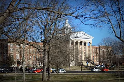 Stately brick building with white columns and a pediment, visible behind bare trees in daylight.