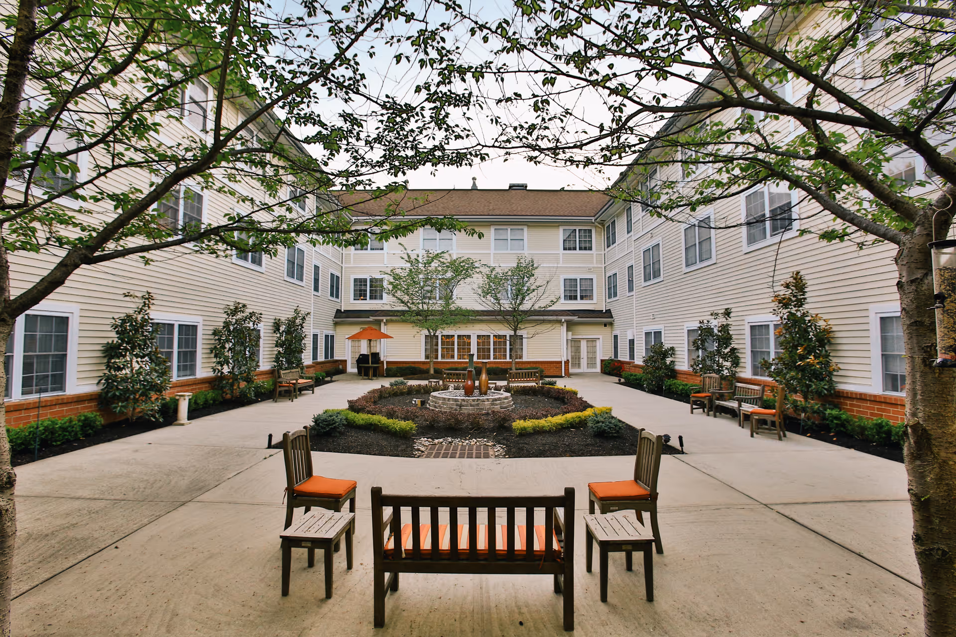 Outdoor courtyard area at Brandywine Pennington by Monarch featuring a central garden with a small fountain, surrounded by benches and chairs with orange cushions. The courtyard is enclosed by a three-story building with multiple windows and some trees framing the view.