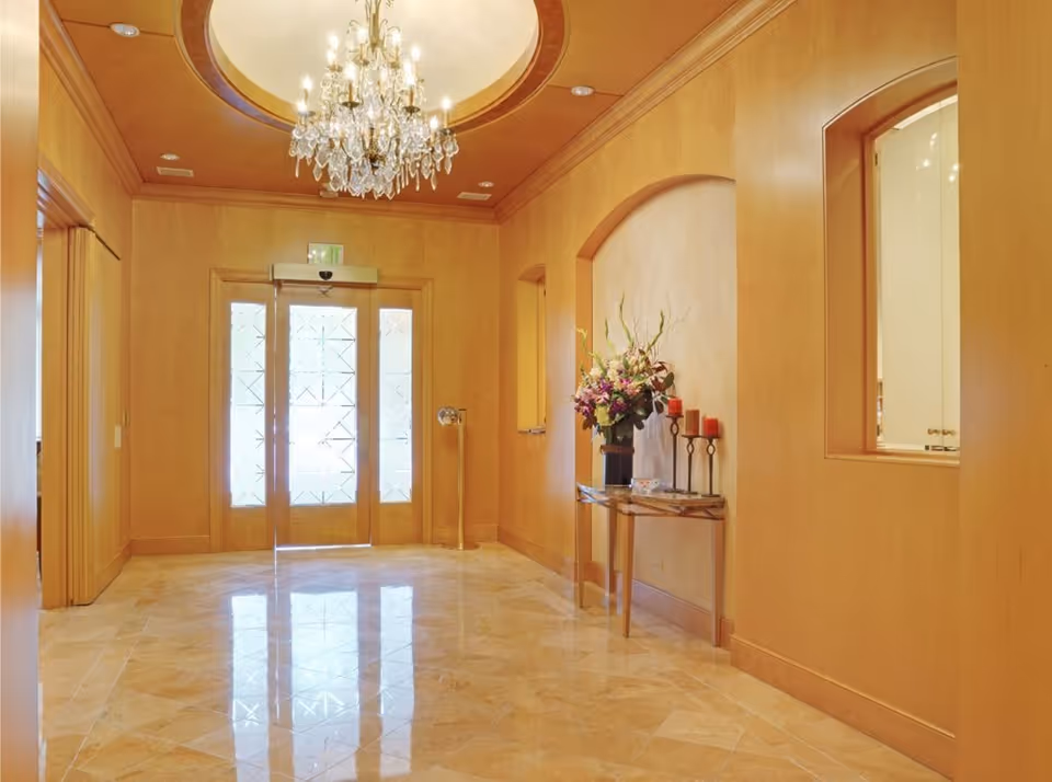 A bright elegant entrance lobby with a chandelier, double glass doors, marble floor and a console table topped with flowers and candles.
