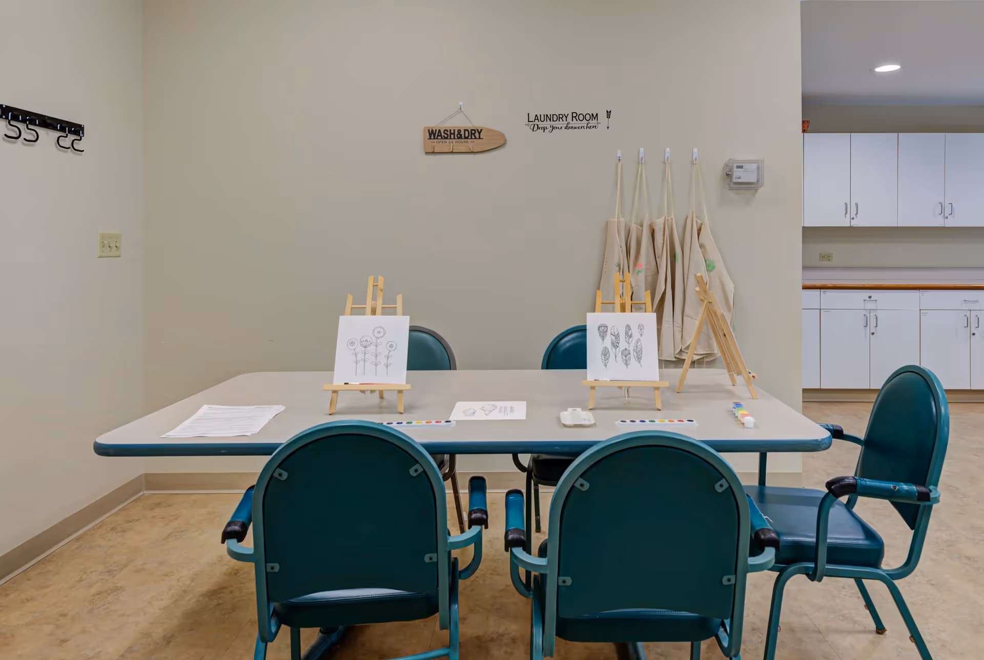 A room with a rectangular table surrounded by four green chairs. On the table are two small easels holding black and white drawings, paint palettes, and paintbrushes. The wall behind the table has a sign that reads 'Laundry Room Drop your drawers here' and several aprons hanging on hooks. White cabinets and countertops are visible in the background.
