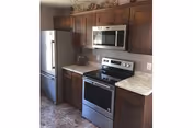 Modern kitchen with stainless steel refrigerator, microwave, and electric stove. The kitchen features dark wooden cabinets and light-colored countertops with a tiled floor.