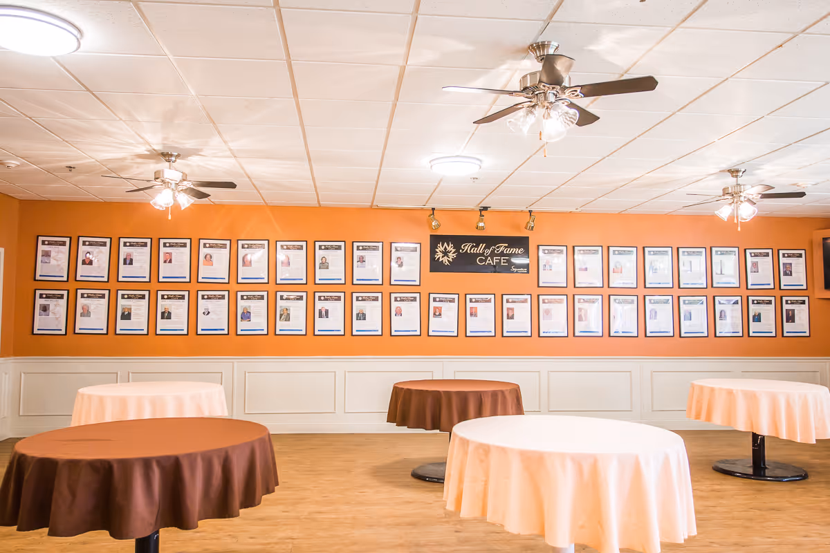 Interior view of a cafe area with round tables covered in brown and white tablecloths. The walls are painted orange with white wainscoting, and numerous framed photos and documents are displayed on the wall. A sign reads 'Hall of Fame Cafe'. Ceiling fans with lights are mounted on the white tiled ceiling.