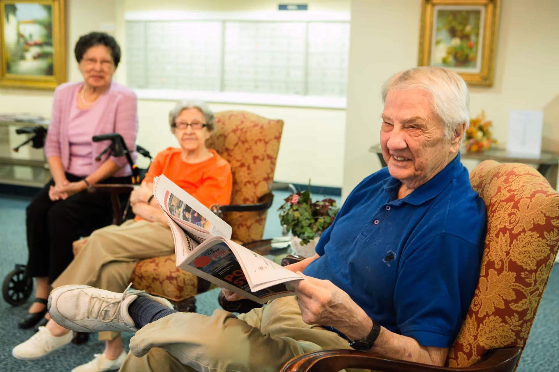 Three elderly residents sitting in a lounge area, a smiling man in the foreground reading a magazine.