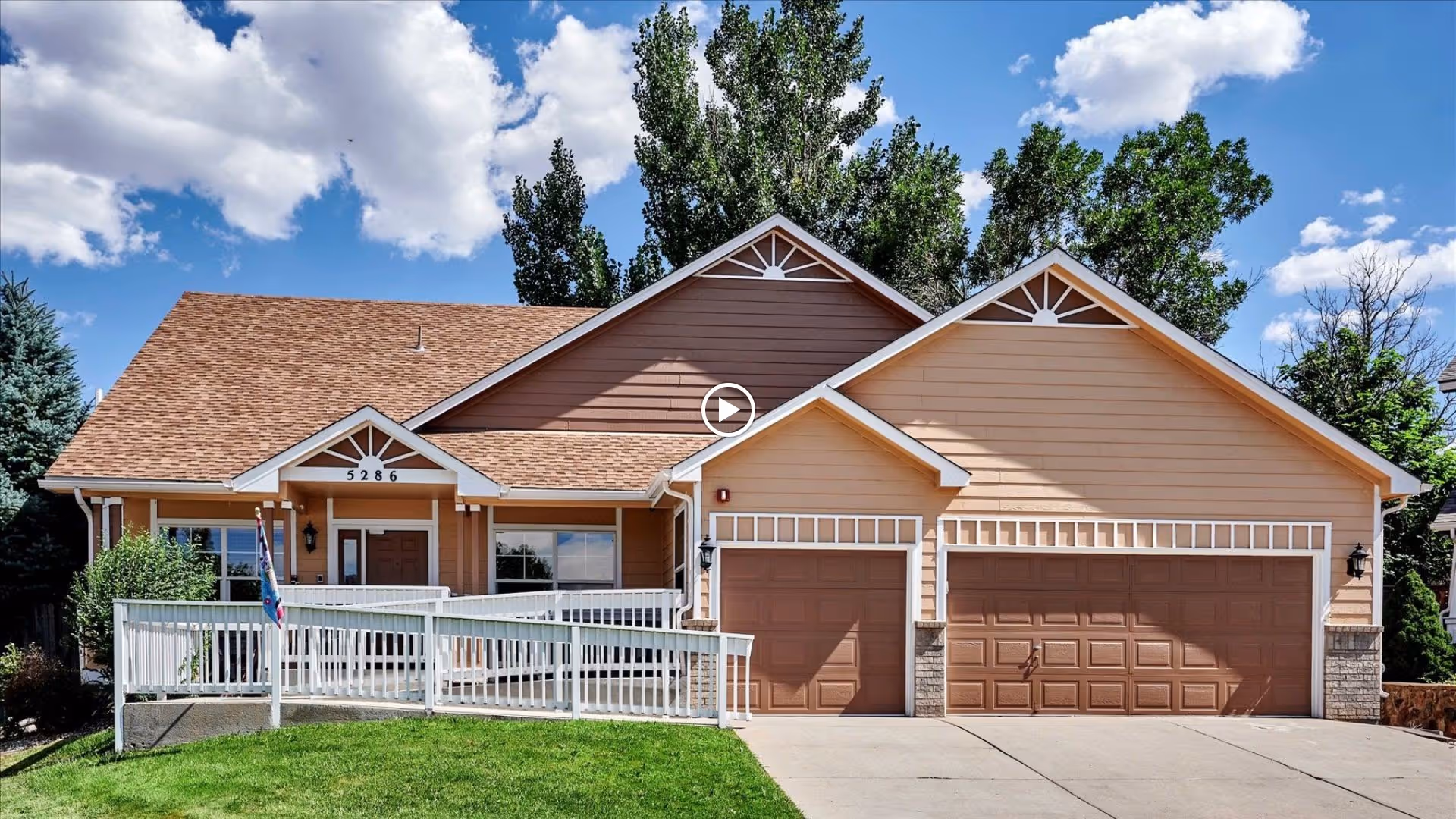 Front exterior of a light-brown single-story home with a porch, ramp, and three-car garage under a blue sky.