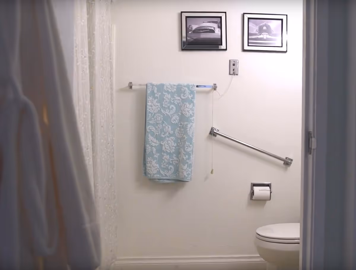 View into a bathroom showing a white toilet, a wall-mounted toilet paper holder, a silver grab bar, a towel rack with a blue and white patterned towel, two framed black and white pictures on the wall, and a white shower curtain partially visible on the left side.