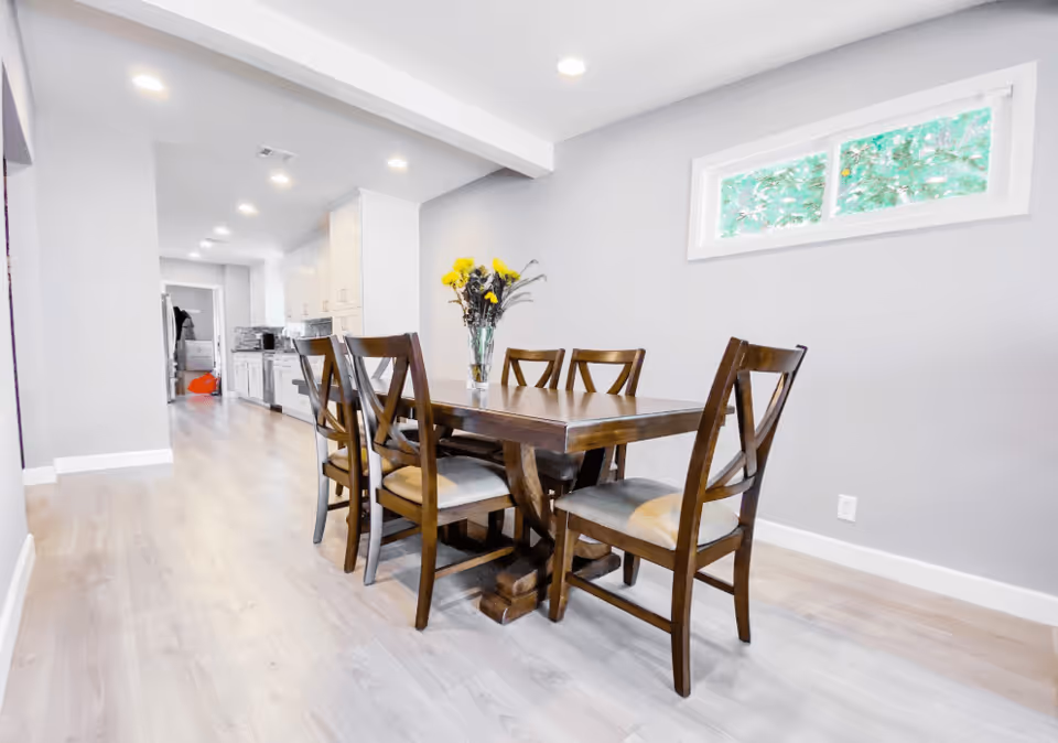 A bright dining area with a wooden dining table and six matching chairs. A vase with yellow flowers is placed on the table. The room has light-colored wooden flooring, gray walls, and a small rectangular window showing greenery outside. The dining area leads into a kitchen with white cabinets and stainless steel appliances.