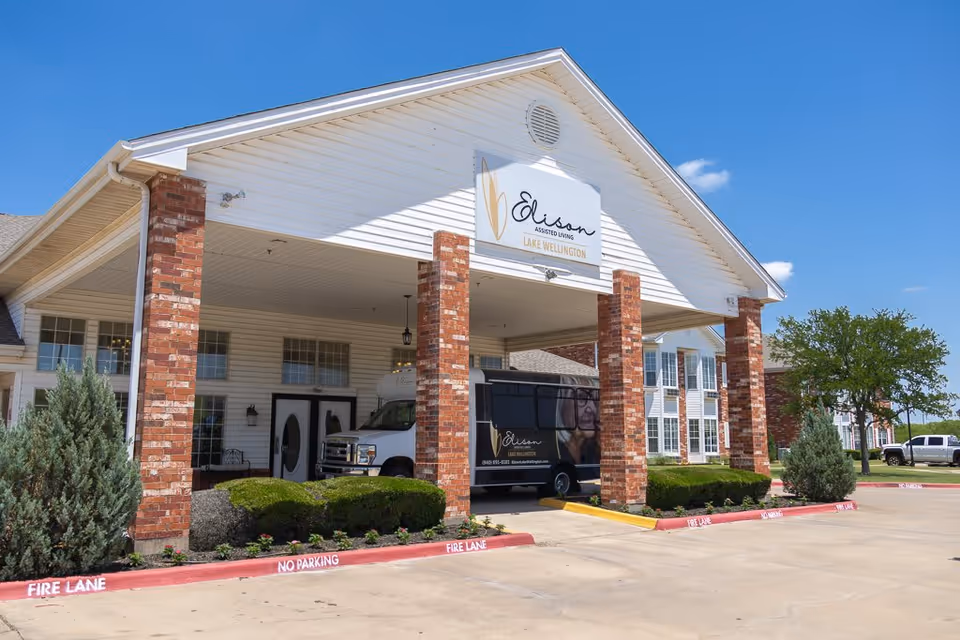 Front exterior view of Elison Assisted Living of Lake Wellington building with a covered entrance supported by brick columns, a parked van with the facility's branding, shrubs, and a clear blue sky.