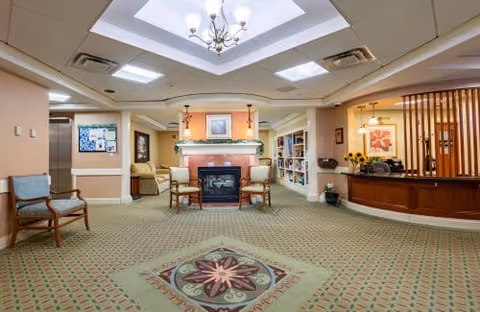A spacious and well-lit common area in a senior living facility featuring a central fireplace with two chairs in front, a patterned carpet, a reception desk to the right, and bookshelves along the back wall. The ceiling has recessed lighting and a chandelier.